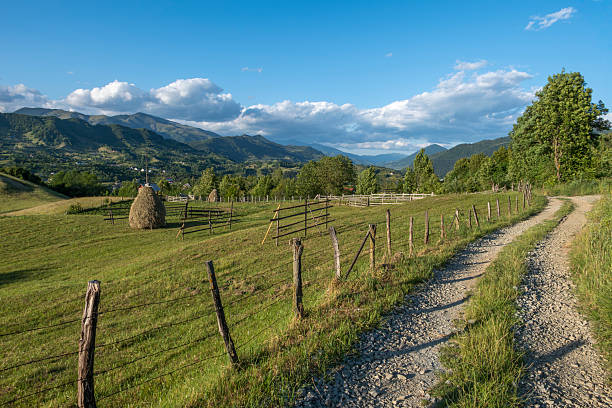 Scenic rural mountain landscape with a dirt road, wooden fence and haystack, surrounded by green meadows and rolling hills under a blue sky, representing traditional countryside life and nature.