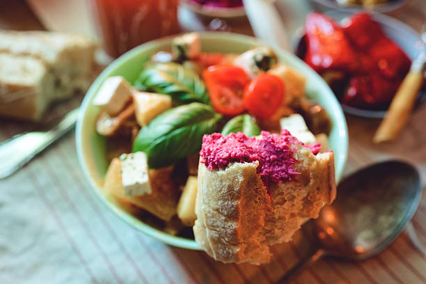 A piece of bread topped with vibrant beet spread held in focus against a softly blurred ratatouille-style dinner table in warm light. The scene offers copy space in the background and along the table surface for editorial text, menu design, and Mediterranean food storytelling. The mood feels cozy, artisanal, and comforting.