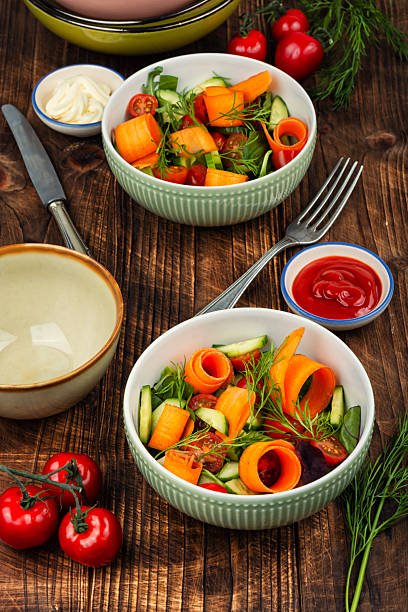 A plate of fresh vegetables on a white background. The plate contains greens, cucumbers, carrots, lemons, and corn. To the right of the plate is another cucumber, a large carrot, and a lemon. All of this is on a white plate.