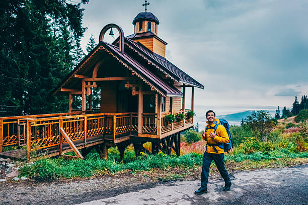 Hiker in a bright yellow jacket and blue backpack walks along a trail near a small mountain chapel surrounded by lush greenery and tall evergreen trees. The rustic chapel is located on a picturesque hill with a narrow mountain path next to it.