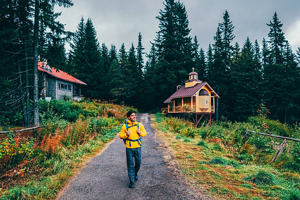 Hiker in a bright yellow jacket and blue backpack walks along a trail near a small mountain chapel surrounded by lush greenery and tall evergreen trees. The rustic chapel is located on a picturesque hill with a narrow mountain path next to it.