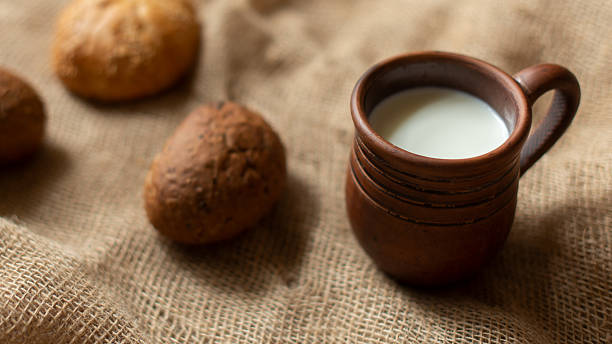 Rustic still life with fresh milk in clay mug and homemade buns on burlap background, selective focus. Rural living, traditional breakfast, handcrafted pottery, food, farm-fresh, cozy, authentic