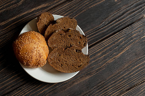 Sesame seed bun and several slices of dark, rustic rye bread on white plate on textured, dark brown wooden table, copy space. Traditional baking, natural ingredients, menu, food, bread basket concept