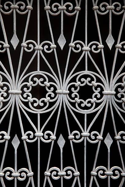 Close-up of a decorative wrought iron pattern featuring symmetrical ornamental shapes and curved metal elements on a dark background. The image works as an architectural detail, abstract texture, or elegant background for design and visual projects.