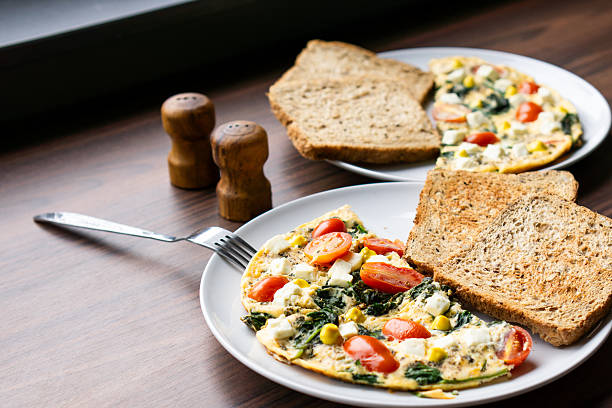 A breakfast plate featuring omelette with spinach, cheese, cherry tomatoes, and corn, accompanied by toast and salt shakers in background. The rustic setting creates a cozy homemade meal atmosphere. Perfect for breakfast and nutrition concepts.
