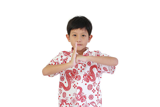 Asian boy wearing traditional white and red dragon-patterned shirt, performing respectful Chinese greeting gesture (Gong Xi) on a white background, celebrating the Chinese New Year festival.