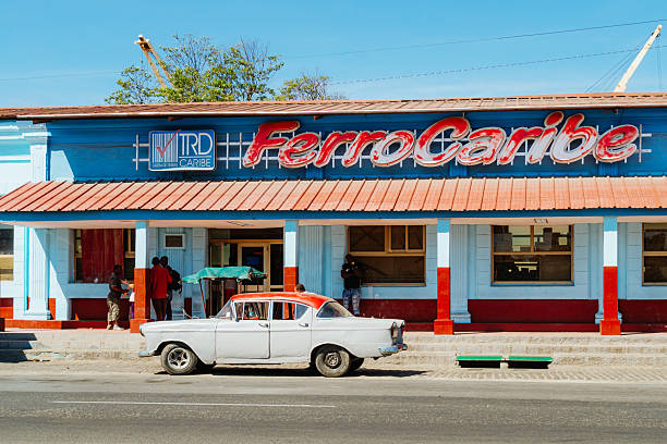 Santiago de Cuba, Cuba on February 15, 2017: A classic white car parked along a vibrant blue Cuban storefront in Santiago de Cuba. Bright red accents and a retro neon sign create a lively, nostalgic street scene.