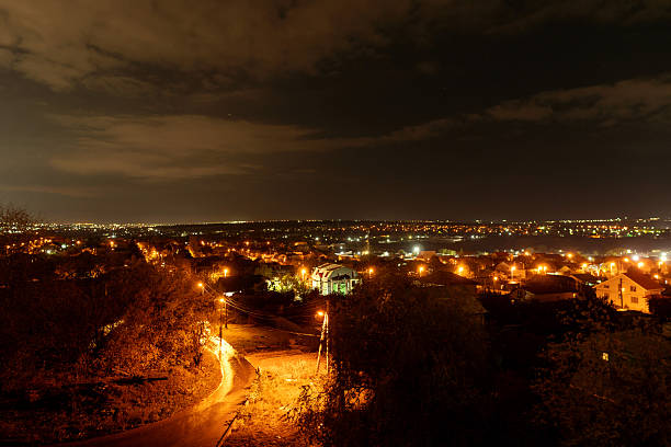 A suburban area at night illuminated by warm street lights, showing houses, trees, and winding roads under a cloudy dark sky with distant city lights on the horizon.