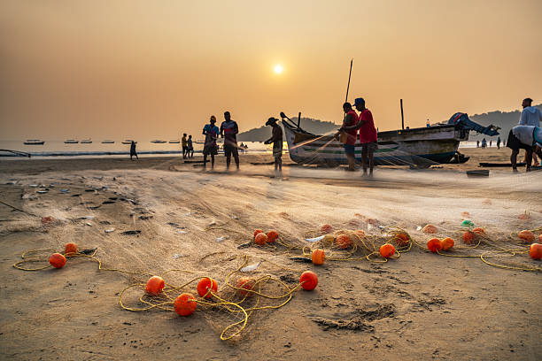 Golden sunset over Goa beach as fishermen mend nets and prepare boats. Orange buoys dot the sand while distant boats float on calm water, capturing coastal life, teamwork, and warm seaside atmosphere.