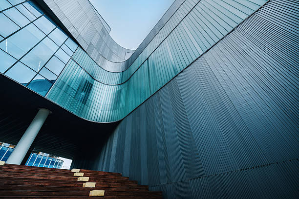 A striking contemporary building features curved glass walls and sleek metal siding. The architectural design emphasizes fluid lines, reflective surfaces, and bold geometry, captured near wooden steps and a supporting column.