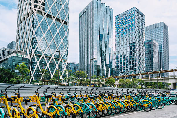 Changsha, China - 28 Feb 2024: A young child in colorful rain gear carries a blue umbrella while navigating a wet plaza in Changsha, framed by towering residential buildings, on a drizzly day. Feb 2024: A man, focused and navigating through traffic, transports multiple large water bottles on his electric bike during a busy day in Changsha, showcasing the everyday hustle of city life.
