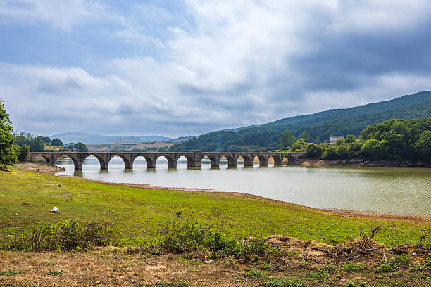 Ancient stone arch bridge crossing the reservoir, surrounded by green hills and cloudy sky