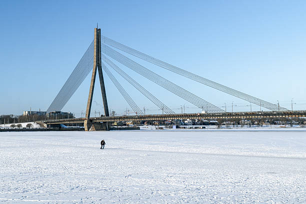 People walking across frozen Daugava River beneath cable-stayed bridge in Riga, illustrating risky winter behavior on unsafe river ice, 02.02.2026. Riga. Latvia