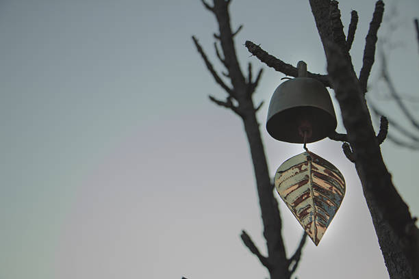 Minimal metal wind bell with leaf ornament hanging on tree branches against soft sky, calm zen atmosphere and peaceful outdoor mood.