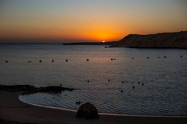 Sun descending creating a vivid orange sky above calm waters and rugged shore