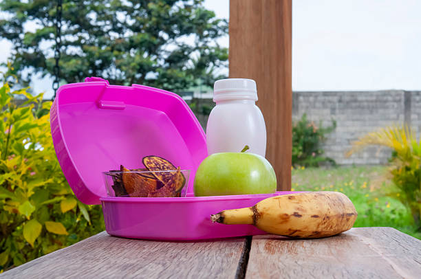 A low-angle shot, taken at table level, shows an open pink lunchbox containing chips, a green apple, a banana, and yogurt on a wooden surface with a blurred garden background.