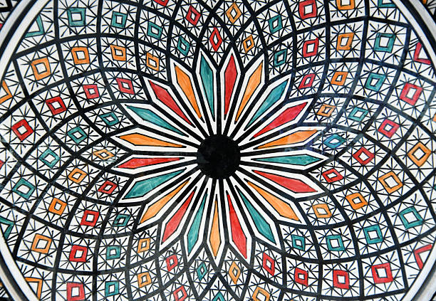 Full-frame close-up view of a traditional ceramic plate originating from Southern Morocco. The earthenware surface features a complex, hand-painted geometric pattern characteristic of Maghrebi and Islamic decorative arts. A central star-shaped motif radiates outward, composed of elongated petal-like shapes filled with red, orange, and turquoise glazes bordered by thick black lines. The background consists of a detailed lattice network containing small squares and diamonds in corresponding colors on a white base. The uneven lines and slight variations in glaze thickness indicate artisanal craftsmanship and the rustic nature of rural Moroccan pottery. The design reflects the cultural heritage of North African geometric ornamentation.