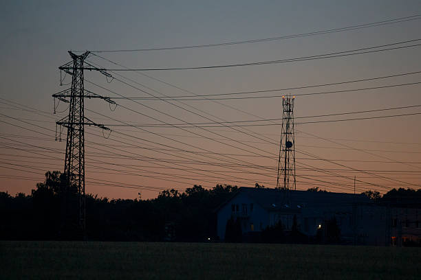 dusk vista of transmission towers and wires, broad scene with multiple pylons crisscrossing above small village buildings, warm dusk glow,