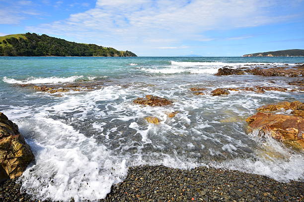 Coastal surf on rocks and shingle beach in Jones Bay with Kawau Island in background. Location: Auckland New Zealand