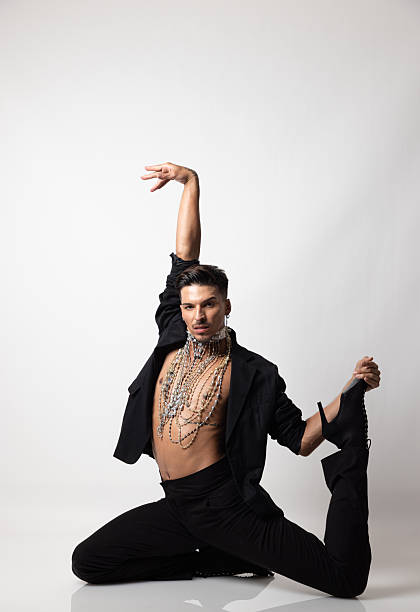 This high-impact studio portrait captures a young man in a dynamic, flexible pose, blending masculine tailoring with elaborate jewelry and expressive makeup. The minimalist white background and clean lighting emphasize the subject's confidence and the intricate details of his styling. This image is perfect for buyers looking to represent modern identity, gender expression, or artistic performance in contemporary lifestyle and fashion campaigns.