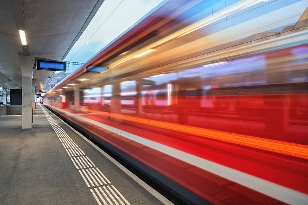 Blurred red passenger train passing mountain railway station in Swiss Alps at night. Moving speed train at dusk. St. Moritz, Switzerland. Bernina Express.  Railway platform and night lights. Transport