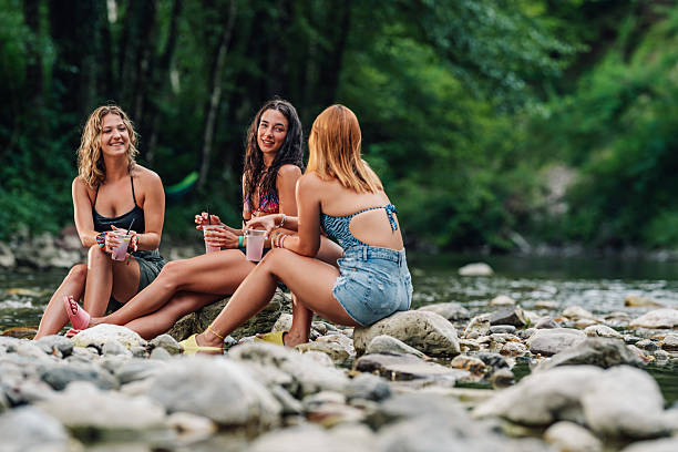 Three young women are sitting on rocks by a river, enjoying refreshing drinks and engaging in lively conversation, surrounded by the beauty of nature
