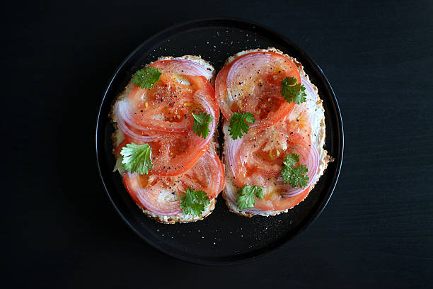 Open sandwich topped with fresh tomato slices, cream cheese, sliced red onion, and herbs, served on a blue plate against a dark background.