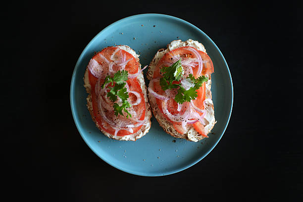 Open sandwich topped with fresh tomato slices, cream cheese, sliced red onion, and herbs, served on a blue plate against a dark background.