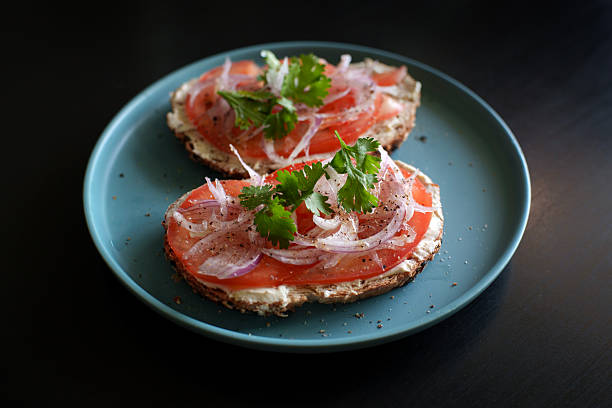 Open sandwich topped with fresh tomato slices, cream cheese, sliced red onion, and herbs, served on a blue plate against a dark background.