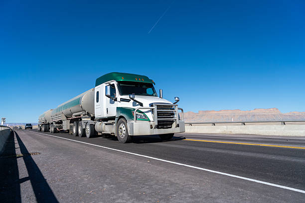 White and green semi truck pulling multiple tanker trailers driving on an empty highway with clear blue sky and desert landscape in background. Large commercial vehicle transporting liquid cargo across open highway with wide lane and minimal traffic.