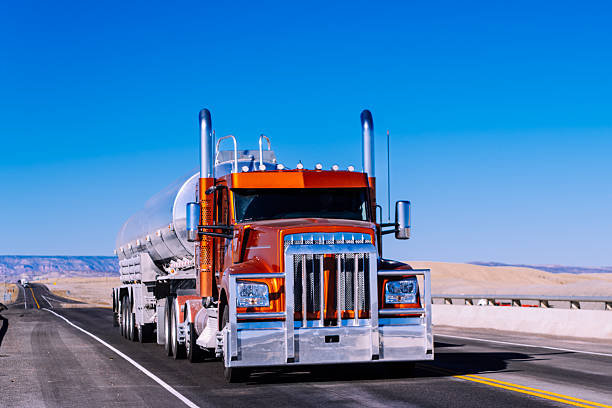 Large orange semi-truck with tanker trailer driving on an empty desert highway under bright blue sky. Vehicle travels through arid landscape with distant mountain ranges, showcasing long-haul transportation infrastructure in an expansive southwestern environment.