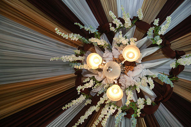 low-angle shot looking directly up at an ornate ceiling decoration, likely for a wedding, banquet, or formal event. At the center is a three-arm chandelier with frosted glass globes emitting a warm, soft glow