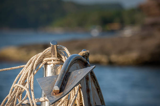 A candid close-up shot of a polished chrome anchor and thick ropes coiled on a boat deck, with blurred rocks and water in the sunny background.
