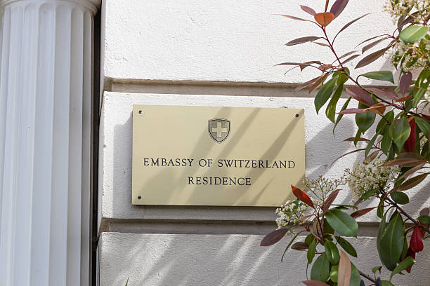 A gold brass plaque reading Embassy of Switzerland Residence mounted on a white wall next to a column and green foliage. London, UK, 14 April 2024