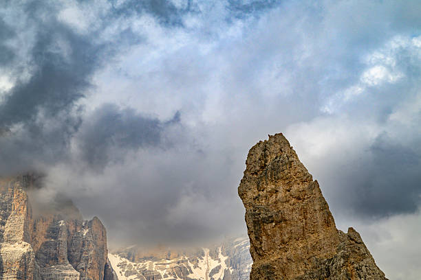 The iconic Cinque Torri (Five Towers) rock formation dominates the landscape in the Dolomites of northern Italy, near Cortina d'Ampezzo. This striking group of limestone towers, set against a dramatic sky with heavy clouds, showcases the rugged beauty of the European Alps during summer. Green alpine meadows and winding trails lead up to the base of these famous peaks, making it a popular destination for hikers, climbers, and photographers. The image captures the majestic scale and unique geological features of this UNESCO World Heritage site. Ideal for travel and tourism promotions, nature documentaries, outdoor adventure content, and editorial pieces on Italian landscapes or European mountain ranges.