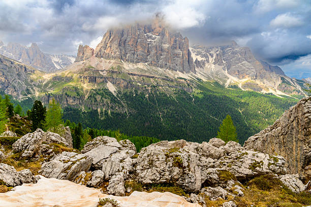 A majestic view of the Tofana di Rozes mountain, part of the Tofana mountain group, in the Italian Dolomites. The rugged peaks rise dramatically under a sky filled with dynamic clouds, some partially obscuring the summit. The foreground features lush green grass and distinctive rocky formations, hinting at the unique geology of the region. This non-urban scene captures the raw beauty and wilderness of the European Alps during a springtime day in Veneto, near Cortina d'Ampezzo. Ideal for travel and tourism promotions, nature and outdoor adventure content, editorial pieces on Italy or the Dolomites, and environmental themes. The absence of people emphasizes the untouched natural landscape.