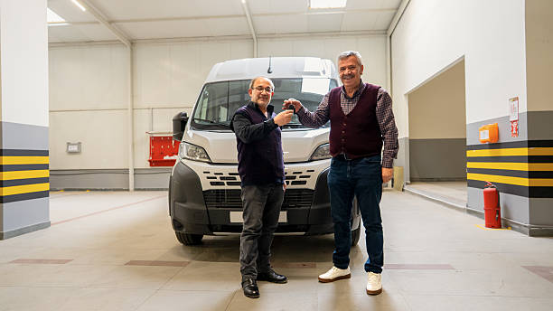 Two adult men smiling while handing over vehicle keys in front of a new panel van inside an automotive workshop. Commercial vehicle delivery, customer satisfaction and business agreement concept.