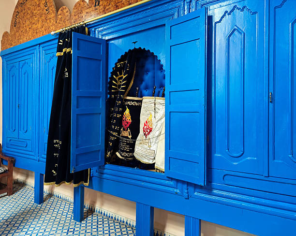 Essaouira / Mogador, Marrakesh-Safi, Morocco: the historic Haim Pinto Synagogue located in the Mellah (Jewish quarter) of Essaouira, Morocco. The image features the central Torah Ark (Hekhal), constructed of wood and painted in a distinct bright blue. The cabinet doors are open, revealing two Torah scrolls encased in embroidered velvet mantles, one black and one cream, adorned with Hebrew calligraphy and iconography. A dark blue velvet curtain (Parokhet) with gold embroidery hangs to the left side of the ark. The floor is paved with traditional blue and white geometric tiles.