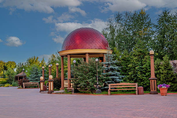 A red dome-shaped gazebo with a yellow base surrounded by trees and benches set against a blue sky with clouds..