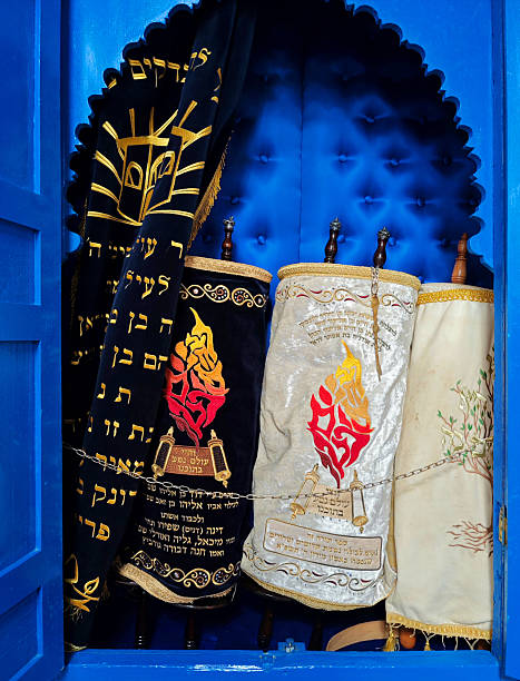 Essaouira / Mogador, Marrakesh-Safi, Morocco: open Aron Kodesh (Holy Ark) at the Rabbi Haim Pinto Synagogue in the Mellah of Essaouira (old Jewish ghetto). Three Sefer Torah scrolls resting upright, encased in velvet Torah Mantles (called Me'il in Hebrew). adorned with embroidered Hebrew calligraphy and stylized flame motifs in gold, red, and silver threads. To the left, a black velvet Parokhet (curtain). The Parokhet features heavy gold embroidery depicting the Tablets of the Law and extensive dedication text. The surrounding wooden cabinetry is painted in a distinctive shade of blue characteristic of the synagogue's design. This site is a significant historical landmark and pilgrimage destination dedicated to the revered tzaddik Rabbi Haim Pinto.