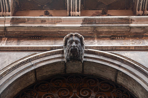 Decorative stone face sculpture integrated into the facade of a historic building in Venice, Italy. The image shows a carved stone head positioned above an arched doorway, framed by weathered architectural elements and aged materials. A close view of traditional Venetian craftsmanship and architectural ornamentation in the historic center.