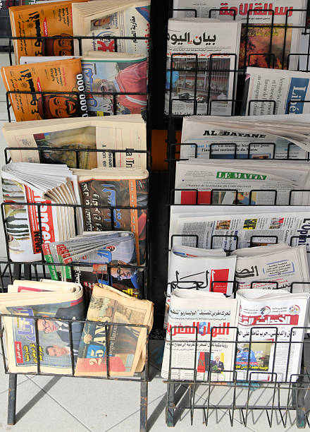 Agadir, Morocco: black wire display rack located in Agadir, Morocco, stocked with a selection of national daily newspapers. The stand features a bilingual mix of publications in Arabic and French, reflecting the linguistic landscape of the country. Visible titles include francophone papers such as Le Matin, Al Bayane, Libération, and L'Opinion, alongside Arabic dailies like Al Ahdath Al Maghribia, Assabah, Bayane Al Yaoum, Rissalat Al Oumma, and Annahar Al Maghribia. Several front pages feature photographs of King Mohammed VI, with headlines addressing national politics, the economy, and regional developments. The papers are folded and stacked in vertical rows within the metal frame, situated outdoors in daylight.