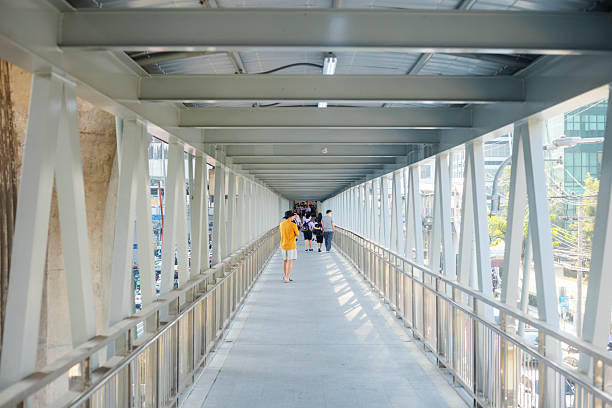 View  along elevated footbridge below BTS  skytrain between  stations  St  Louis and Sarusak at N Sathon Road at afternoon  sunshine. Footbridge  is over road in  center  of the street and leading  from  east to west connected to footbridge  crossing  the road near Pramuan  Road in Silom. Thai people and  students are walking on  footbridge in  far background.   Footbridge is leading to station  Sarusak