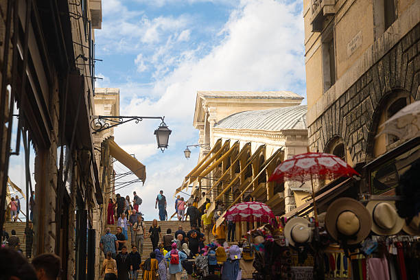 Pedestrians walking along a narrow street near the Rialto Bridge in Venice, Italy. The image shows historic buildings, small shops, and market stalls leading toward one of the city’s most famous landmarks. The scene represents everyday urban life, tourism, and the architectural character of Venice’s historic center.