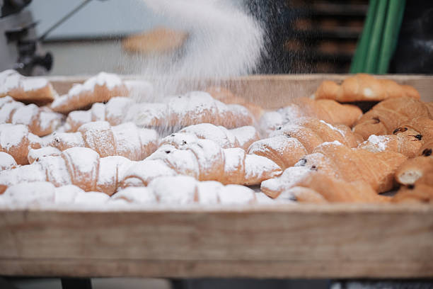 Freshly baked croissants receiving a dusting of powdered sugar on a wooden tray