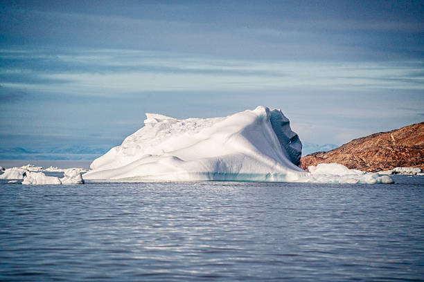 Iceberg floating in Qalerallit Sermia fjord, South Greenland, with the Islandis glacier in the background. The iceberg is a striking blue, with jagged and smooth surfaces, surrounded by floating ice pieces. The fjord s icy waters reflect the pale light. Distant mountainous terrain is obscured by mist, creating a serene, cold atmosphere characteristic of the Arctic landscape.