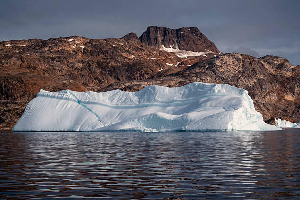 Close-up view of a massive glacier terminus meeting the ocean in East Greenland. Thick fog and mist shroud the upper ice and rocky cliffs, highlighting the deep blue crevasses and jagged texture of the frozen landscape.
