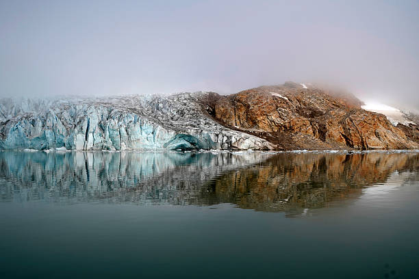 Close-up view of a massive glacier terminus meeting the ocean in East Greenland. Thick fog and mist shroud the upper ice and rocky cliffs, highlighting the deep blue crevasses and jagged texture of the frozen landscape.