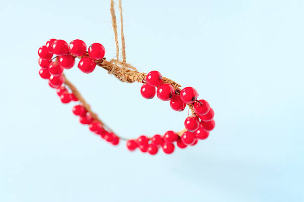 Minimal lifestyle image of a handmade heart shaped decorative ornament crafted from red berry elements and natural twine, hanging against a soft light blue background. The composition feels airy, calm, and romantic, with generous copy space surrounding the heart, making it ideal for Valentine’s Day or Mother’s Day messages, greeting cards, or seasonal marketing.