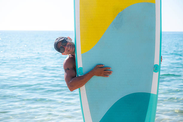 Man smiling and peeking from behind a stand up paddleboard at the beach